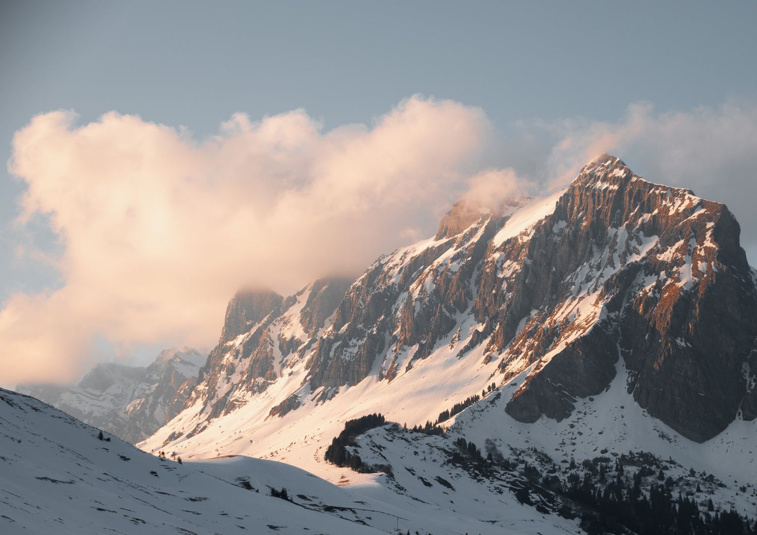 A majestic snow-covered mountain range bathed in soft, golden light from the setting sun, with clouds partially obscuring the peaks against a clear blue sky.